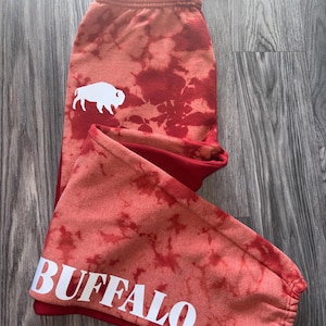 Pode incluir: Calças de moletom vermelhas e laranja com tie-dye, um gráfico de búfalo branco e a palavra "BUFFALO" em letras brancas. As calças de moletom têm cós com cordão e punhos nas tornozelos.