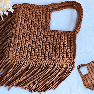 May include: A brown crocheted handbag featuring a square body with a cutout handle and long, flowing fringe trim along the bottom and side edges. The bag is constructed with a textured, ribbed stitch pattern and includes a smaller matching version.