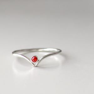 May include: A silver ring featuring a V-shaped design, with a small, round, red gemstone set in the centre. The ring is displayed on a white surface, with blurred decorative objects in the background.