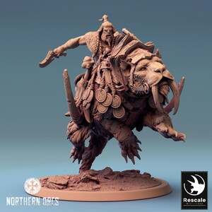May include: A monochromatic brown resin miniature figurine depicting an orc warrior riding a large saber-toothed beast. The orc holds a blade, while the beast wears armor and hanging circular medallions. The base features rocky terrain details.
