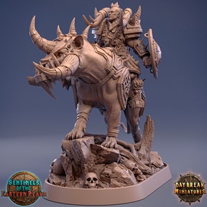 Op de afbeelding: Een miniatuurfiguur van een krijger die een monstrueus beest berijdt. De krijger draagt ​​een harnas en zwaait met een schild. Het beest heeft scherpe tanden en klauwen. Het figuur is geschilderd in een bruin en grijs kleurenschema. De basis van het figuur is versierd met schedels en rotsen. Het figuur is gemaakt door Daybreak Miniatures.