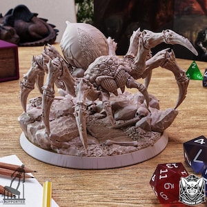 May include: A detailed, gray spider figurine with multiple legs and a large abdomen, posed on a rocky base. The figurine includes small skulls and bones, suggesting a fantasy or tabletop gaming theme. Dice and pencils are visible in the background.