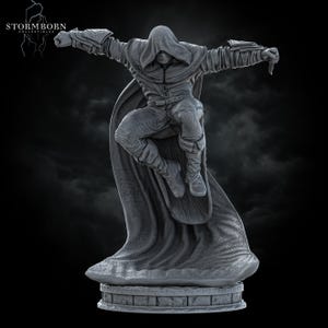 Op de afbeelding: Een grijze beeldje van een figuur met capuchon in een dynamische pose, armen uitgestrekt, met een klein mes. De figuur draagt een cape en gedetailleerde kleding. De basis is een steenachtige structuur. Het Stormborn Collectibles-logo staat linksboven.