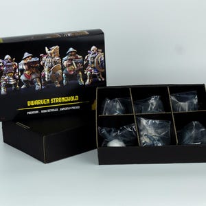 Könnte beinhalten: Ein schwarzes Box-Set mit der Aufschrift "Dwarven Stronghold" mit Miniaturfiguren. Die Box ist geöffnet und zeigt Fächer mit verpackten Figuren. Das Box-Art zeigt detaillierte Illustrationen der Zwergenfiguren.