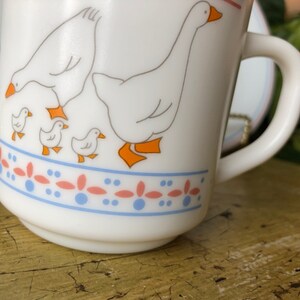 May include: White ceramic mug with a blue and pink border and a design of five white geese. The geese are in a line with the largest goose in the back and four smaller geese in front of it. The geese are all facing the right side of the mug.
