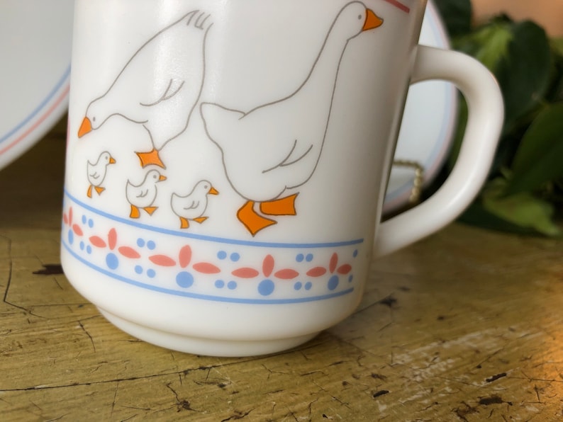 May include: White ceramic mug with a blue and pink border and a design of five white geese. The geese are in a line with the largest goose in the back and four smaller geese in front of it. The geese are all facing the right side of the mug.