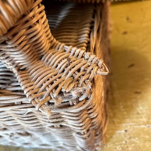 May include: Close-up of a woven wicker basket with a rectangular shape. The basket is made of light brown wicker and has a textured surface. The weave is visible, and some strands are frayed.