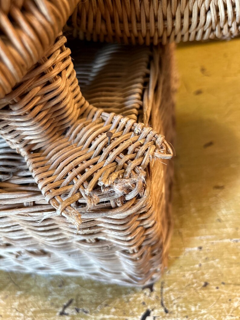 May include: Close-up of a woven wicker basket with a rectangular shape. The basket is made of light brown wicker and has a textured surface. The weave is visible, and some strands are frayed.