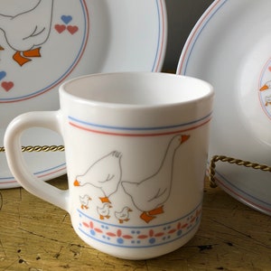 May include: White ceramic mug with a blue and red stripe around the top and a design of geese and goslings on the front. The mug has a white handle.