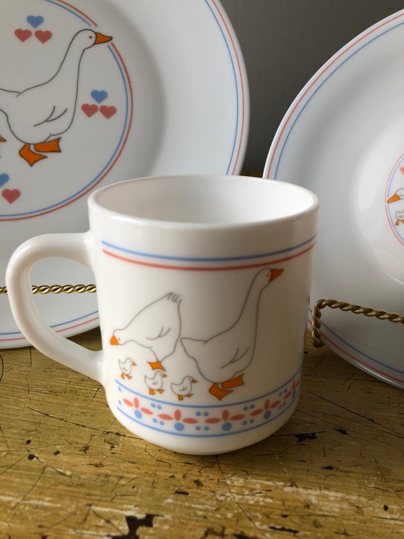 May include: White ceramic mug with a blue and red stripe around the top and a design of geese and goslings on the front. The mug has a white handle.