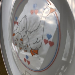 May include: White ceramic plate with a blue, red, and orange border. The center of the plate features a white goose with three goslings. The goose and goslings are surrounded by pink and blue hearts.