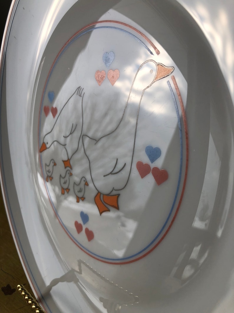 May include: White ceramic plate with a blue, red, and orange border. The center of the plate features a white goose with three goslings. The goose and goslings are surrounded by pink and blue hearts.