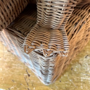 May include: Close-up of a woven wicker basket with a rounded corner. The basket is made of light brown wicker and has a textured surface. The basket appears to be sturdy and well-crafted.