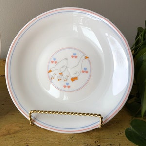 May include: White ceramic plate with a blue and pink border. The center of the plate features a design of a family of geese with orange beaks and feet. The geese are surrounded by pink and blue hearts.