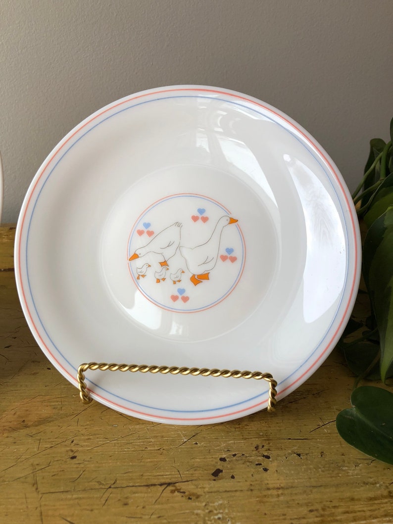 May include: White ceramic plate with a blue and pink border. The center of the plate features a design of a family of geese with orange beaks and feet. The geese are surrounded by pink and blue hearts.