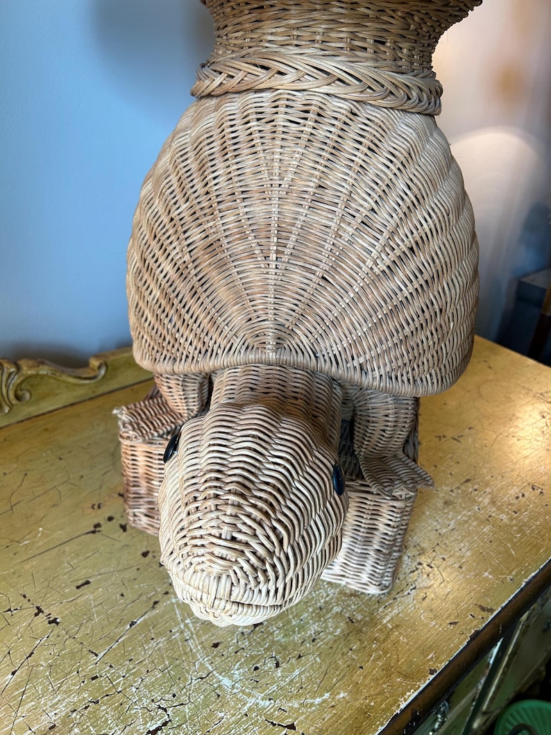 May include: A woven wicker turtle-shaped basket with a textured surface. The turtle has a rounded shell, a detailed head with black eyes, and four legs. The basket is light brown and appears to be handmade.