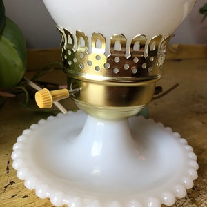 May include: White milk glass table lamp with a gold metal base and a beaded edge on the base. The lamp has a white glass shade with a gold metal trim.