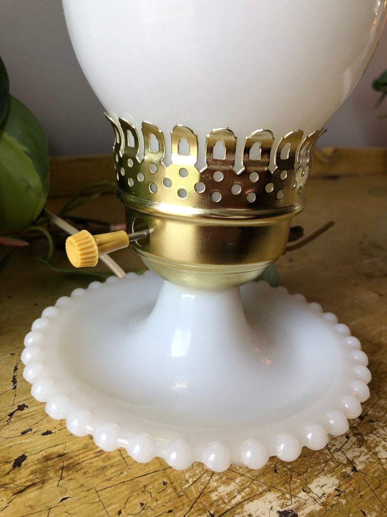 May include: White milk glass table lamp with a gold metal base and a beaded edge on the base. The lamp has a white glass shade with a gold metal trim.