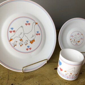 May include: A white ceramic dinnerware set with a blue and red trim. The set features a design of a goose family with hearts. The set includes a dinner plate, a salad plate, and a mug.