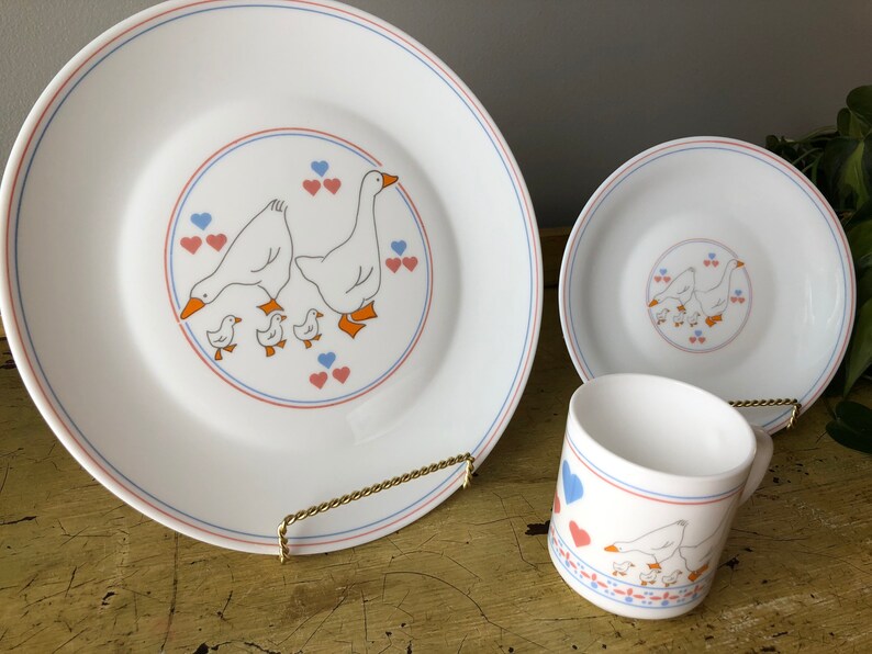 May include: A white ceramic dinnerware set with a blue and red trim. The set features a design of a goose family with hearts. The set includes a dinner plate, a salad plate, and a mug.