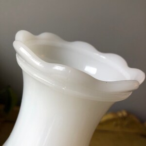 May include: White milk glass vase with a scalloped rim. The vase has a flared opening and a narrow base.