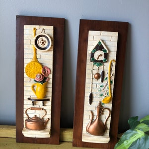 Op de afbeelding: Twee vintage wandplaques met een keukenthema. De plaque aan de linkerkant heeft een gele theepot, een gele kan met roze bloemen en een koperen waterkoker. De plaque aan de rechterkant heeft een koperen theepot, een gele soeplepel en een koekoeksklok.
