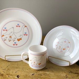 May include: A set of white Corelle dinnerware with a goose and gosling design. The set includes a large plate, a smaller plate, and a mug. The plates have blue and pink trim and the mug has a blue and red trim.
