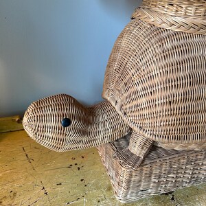 May include: Wicker turtle-shaped storage basket. The basket is made of woven light brown wicker and features a turtle head with a black eye. The turtle's shell forms the main body of the basket, with a lid on top. The basket sits on a yellow surface.