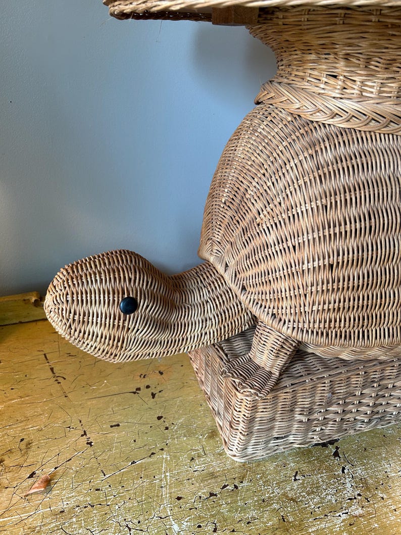 May include: Wicker turtle-shaped storage basket. The basket is made of woven light brown wicker and features a turtle head with a black eye. The turtle's shell forms the main body of the basket, with a lid on top. The basket sits on a yellow surface.