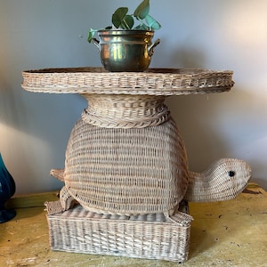 May include: A whimsical, light brown wicker turtle-shaped side table. The table features a round top, a turtle body, and a rectangular base. A brass pot with greenery sits on top. The table is a unique decorative piece.