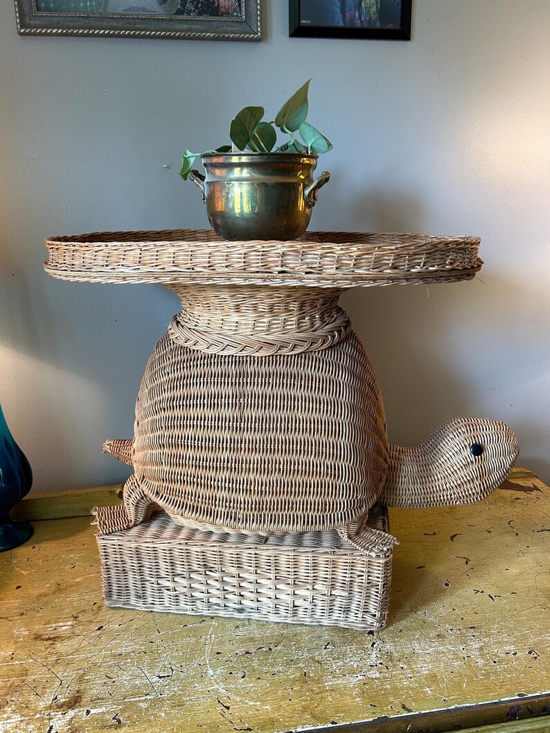 May include: A whimsical, light brown wicker turtle-shaped side table. The table features a round top, a turtle body, and a rectangular base. A brass pot with greenery sits on top. The table is a unique decorative piece.