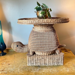 May include: A whimsical, light brown wicker turtle-shaped side table. The table features a round top, a turtle-shaped body, and a rectangular base. A small brass pot with a green plant sits on the table's surface.