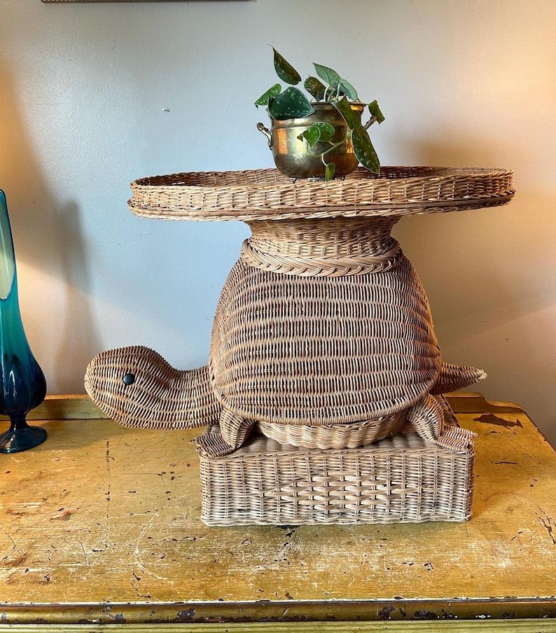May include: A whimsical, light brown wicker turtle-shaped side table. The table features a round top, a turtle-shaped body, and a rectangular base. A small brass pot with a green plant sits on the table's surface.