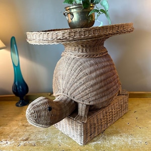 May include: A woven wicker turtle-shaped plant stand. The turtle has a round body, a head, and a rectangular base. A brass pot with a green plant sits on top of the turtle's back. The stand is light brown in color.