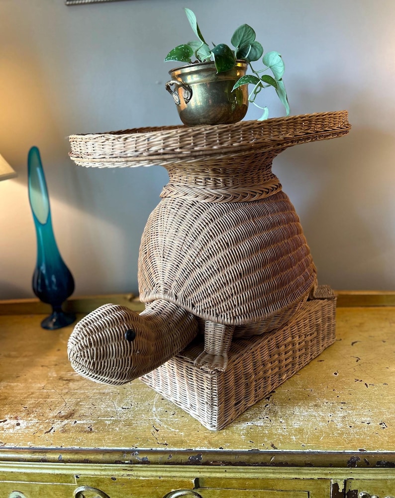May include: A woven wicker turtle-shaped plant stand. The turtle has a round body, a head, and a rectangular base. A brass pot with a green plant sits on top of the turtle's back. The stand is light brown in color.