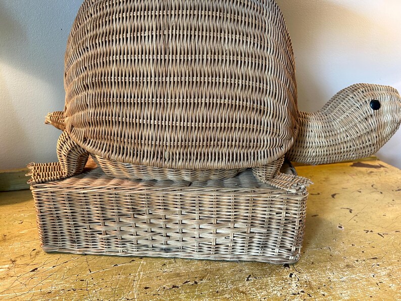 May include: A woven wicker turtle-shaped basket with a rectangular base. The turtle has a rounded shell, a head with a black eye, and four small feet. The basket is light brown and appears to be handmade.