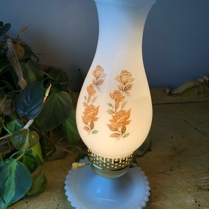 May include: A white milk glass lamp with a scalloped top and a floral design. The lamp has a gold base and a white milk glass base with a beaded edge.