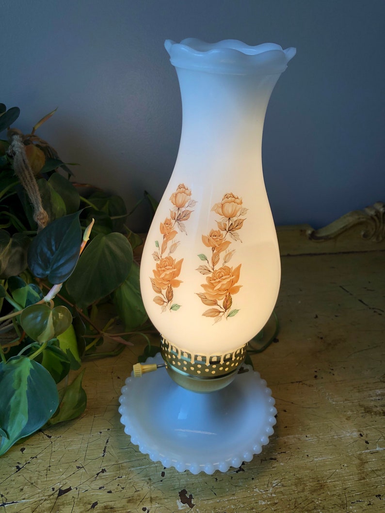 May include: A white milk glass lamp with a scalloped top and a floral design. The lamp has a gold base and a white milk glass base with a beaded edge.