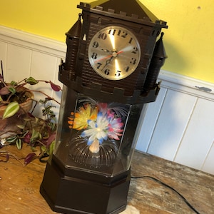 May include: A dark brown novelty clock in a castle design. The clock face is silver with black numerals and red hands. The base has a clear, hexagonal section with colourful fibre optic flowers. A power cord extends from the base.