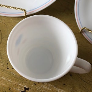 May include: White ceramic coffee cup with a blue and red rim. The cup is empty and sitting on a wooden surface.