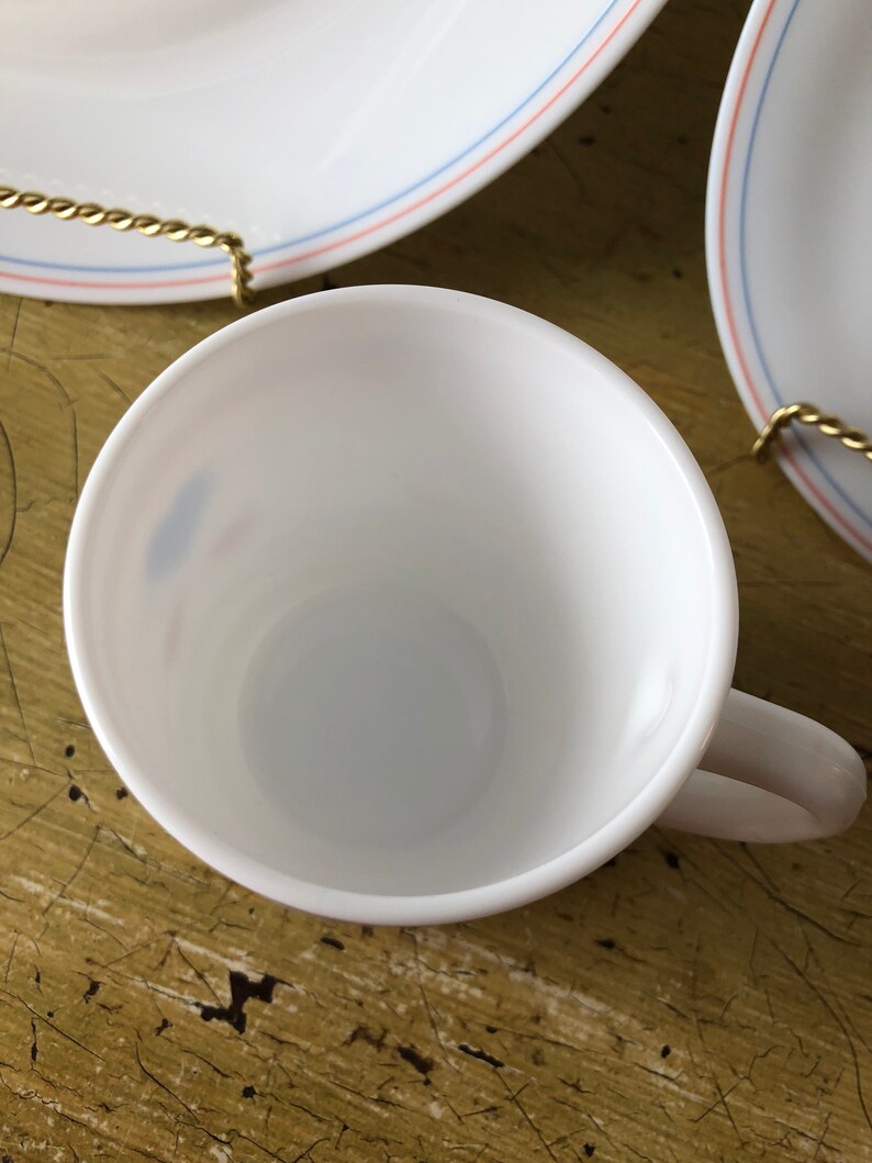 May include: White ceramic coffee cup with a blue and red rim. The cup is empty and sitting on a wooden surface.