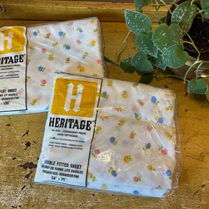 May include: Two packages of Heritage brand bed sheets in white with a small floral pattern in yellow, pink, and blue. One package is a double flat sheet measuring 81 inches by 100 inches, and the other is a double fitted sheet measuring 54 by 75 inches.