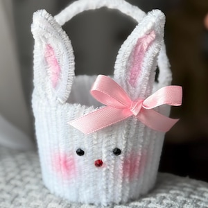 May include: A white bunny-shaped basket with a handle, pink inner ears, and a pink bow. The basket has black eyes and a red nose. The basket is made of a soft, textured material, perfect for Easter or spring decor.