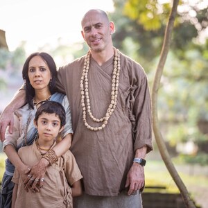 May include: A family of three poses outdoors. The man wears a brown tunic and a beaded necklace. The woman wears a light blue top and a shawl. The child wears a brown tunic and a beaded bracelet. The background is a green, natural setting.