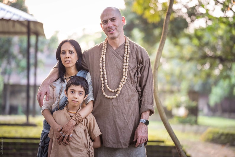 May include: A family of three poses outdoors. The man wears a brown tunic and a beaded necklace. The woman wears a light blue top and a shawl. The child wears a brown tunic and a beaded bracelet. The background is a green, natural setting.