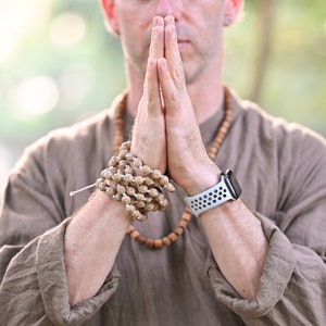 May include: A person wearing a brown shirt and a wooden bead necklace, with hands clasped in prayer. A beaded bracelet and a smartwatch with a white band are visible. The background is blurred with green foliage.