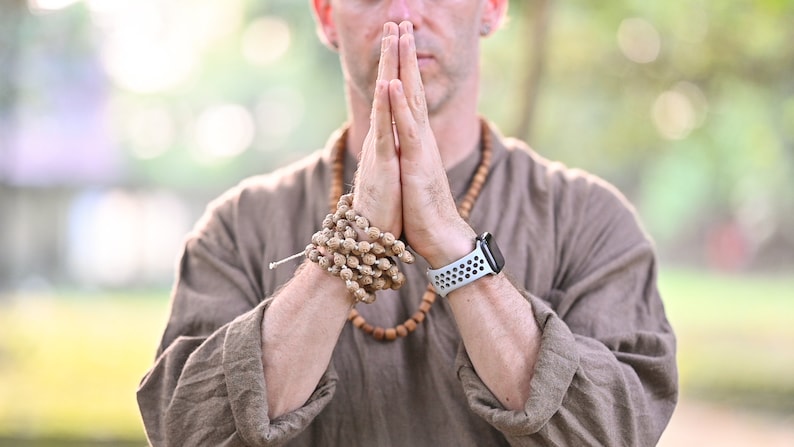 May include: A person wearing a brown shirt and a wooden bead necklace, with hands clasped in prayer. A beaded bracelet and a smartwatch with a white band are visible. The background is blurred with green foliage.