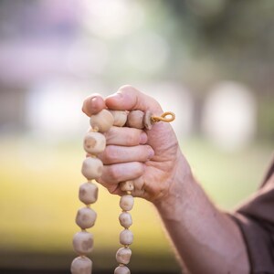 May include: A strand of light-colored wooden prayer beads held in a hand. The beads are round and connected by a string, with a loop at the end. The background is blurred, suggesting an outdoor setting.