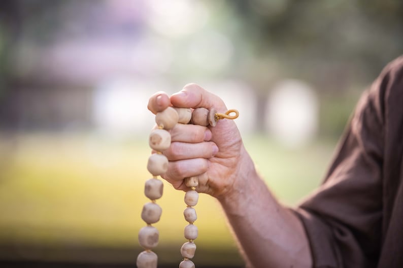 May include: A strand of light-colored wooden prayer beads held in a hand. The beads are round and connected by a string, with a loop at the end. The background is blurred, suggesting an outdoor setting.