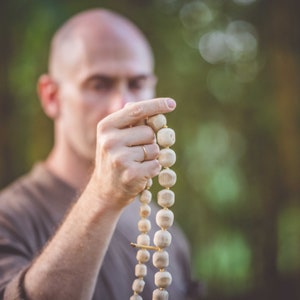 May include: A wooden mala bead necklace with large, rounded beads. The beads are strung together with a gold-colored spacer. The background is blurred, with a person holding the necklace in focus.